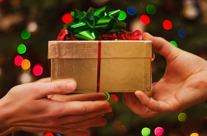 two hands holding a wrapped present in front of a lit Christmas tree